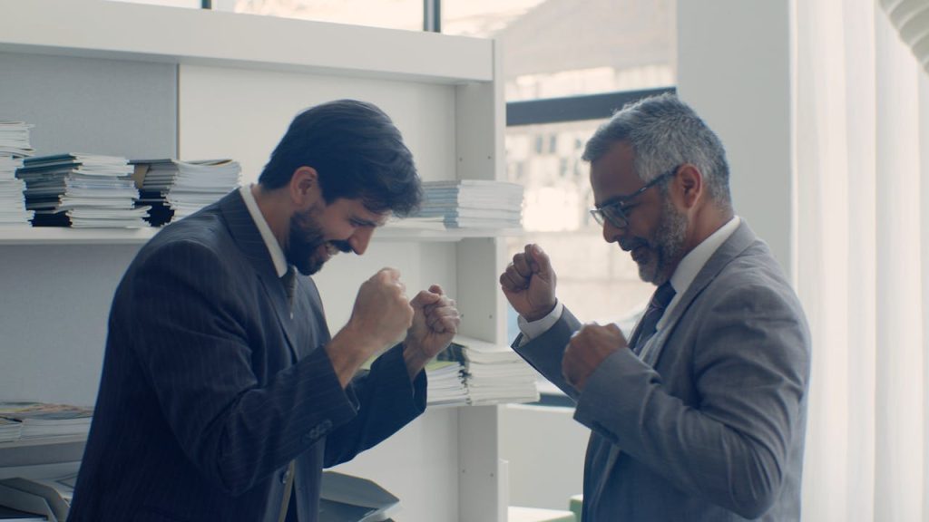 Two businessmen in suits celebrating success with fists raised in a modern office setting.