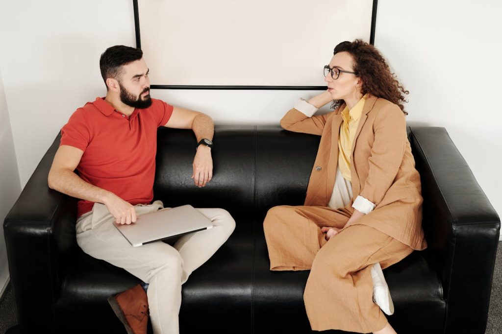 A man and woman sit on a black sofa, having a professional conversation.